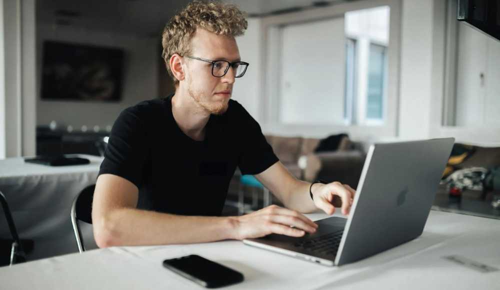 Person with curly hair and glasses using MacBook at a table, iPhone nearby, in a bright room with a couch in the background.