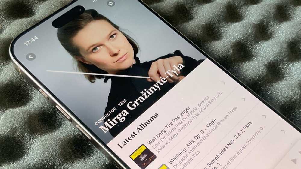 Smartphone displays a classical music conductor with a baton, featuring their name and latest albums on a music app interface.
