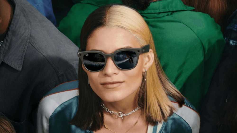 Person with sunglasses and two-tone hair, wearing a patterned jacket and necklace. Sunglasses reflect outdoor scenery with clouds and sky.