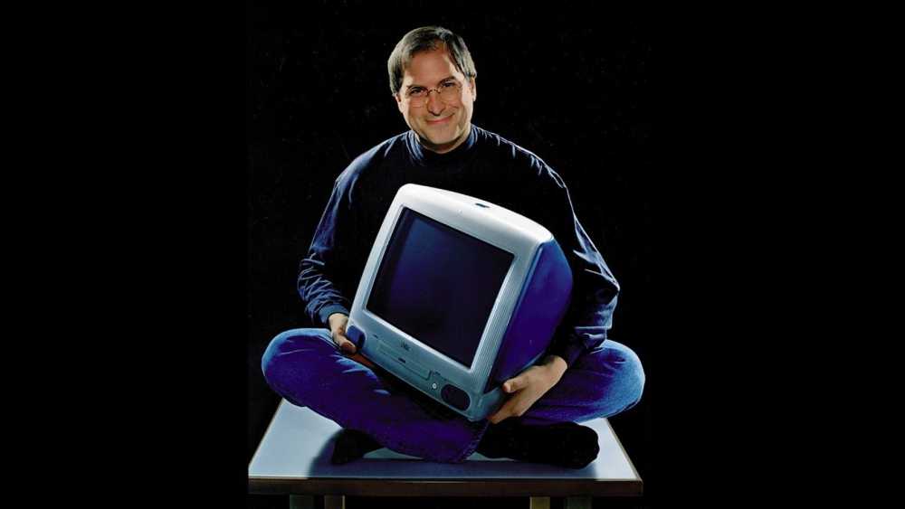 Person with glasses, smiling, sitting cross-legged, holding an old Apple iMac with a translucent case, against a dark background.