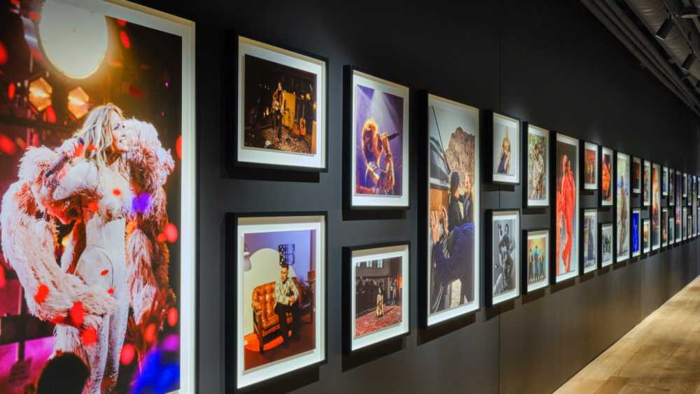 A gallery wall displaying various framed photographs, predominantly featuring musicians performing and candid moments captured in vibrant colors, under dim warm lighting.
