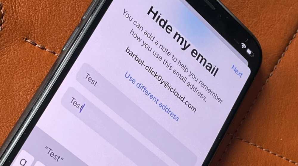 Smartphone screen displaying the 'Hide my email' feature with a test email and note entry fields, on a brown leather surface.