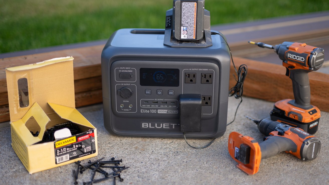 Portable power station with attached cable on concrete, accompanied by a yellow box of screws, and two orange cordless drills on the ground. Grass is visible in the background.