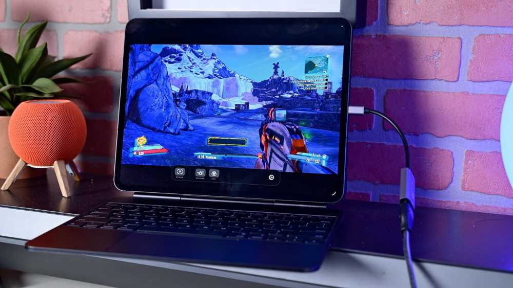 Tablet displaying a snowy video game scene with a shooter perspective, keyboard below, orange speaker on the left, and a charger plugged in on the right.