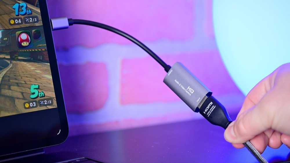 Hand holding HDMI cable connected to an audio-video capture device near a laptop screen showing a racing game. Background features a pink and blue gradient.