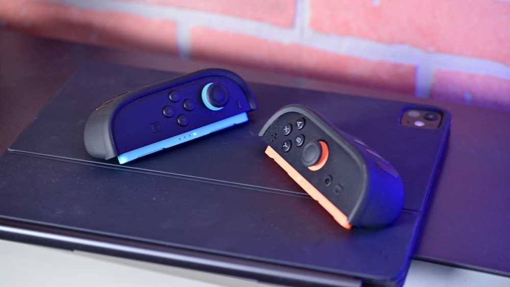 A hand places a tablet into a docking station beside books and a colorful model tripod on a white shelf with a brick wall background.