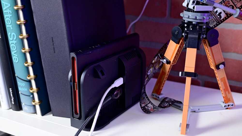 Books on a shelf beside a gaming console dock, with a connected cable and a small LEGO tripod in the background.