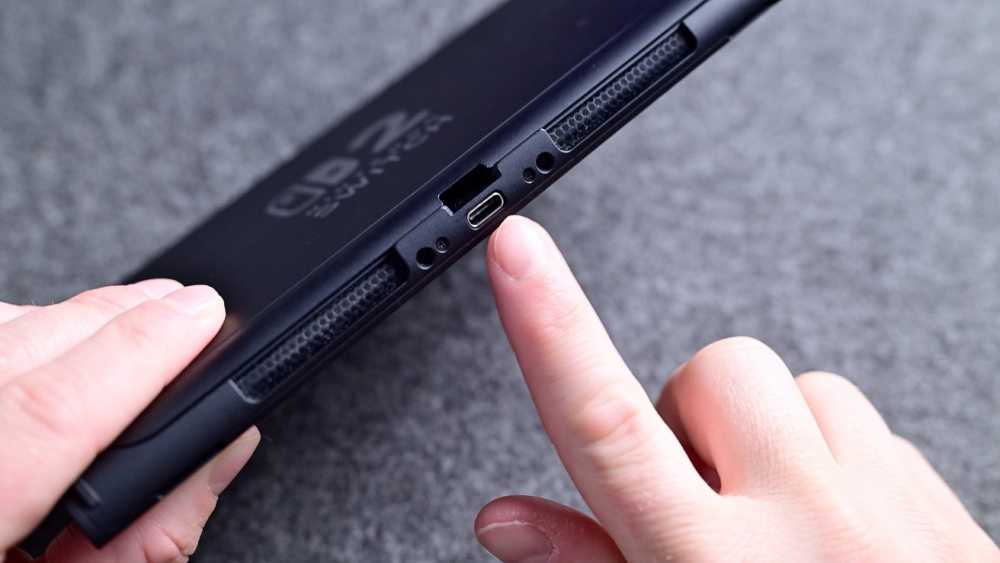 Hands holding and pointing at a black electronic device with a USB-C port on a gray textured surface.