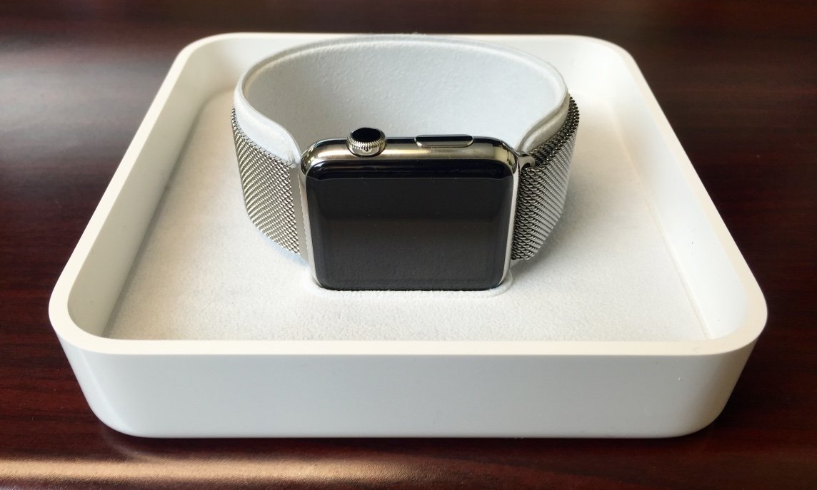 A smartwatch with a dark screen and a silver mesh band sits on a white square display tray.