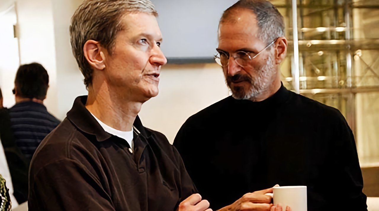 Two men in conversation, one holding a white mug, standing indoors with blurred background.
