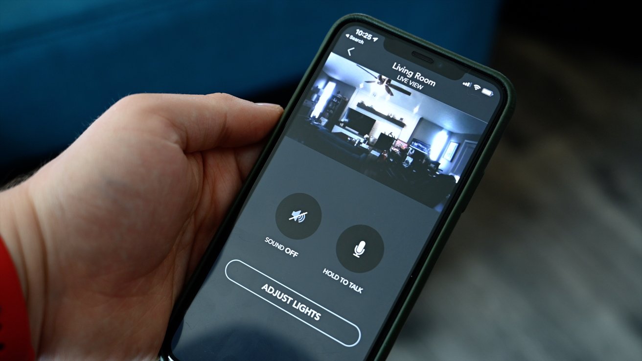 Hand holding a smartphone displaying a live view of a living room, with options to mute sound, talk, and adjust lights.