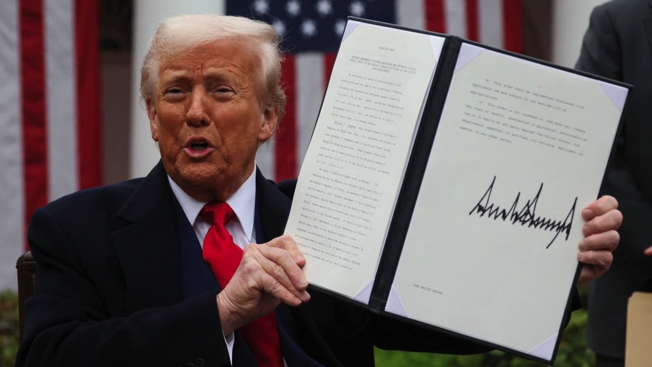 Man with light hair and black suit holding an open folder showing a signed document. United States flag in the background.