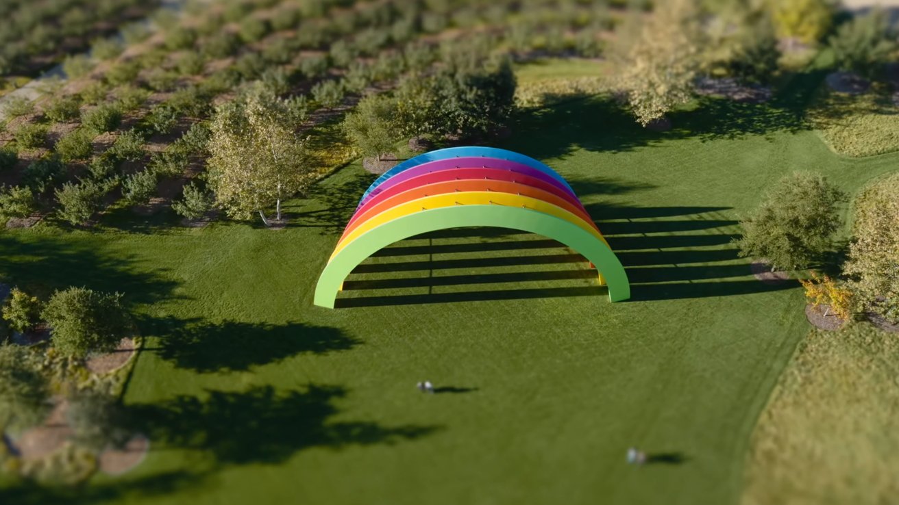 A colorful rainbow arch sculpture stands on a large grassy field surrounded by trees, casting long shadows.