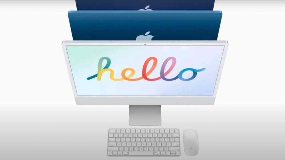 White desktop computer with colorful 'hello' on the screen, Apple logo behind, accompanied by a keyboard and mouse.