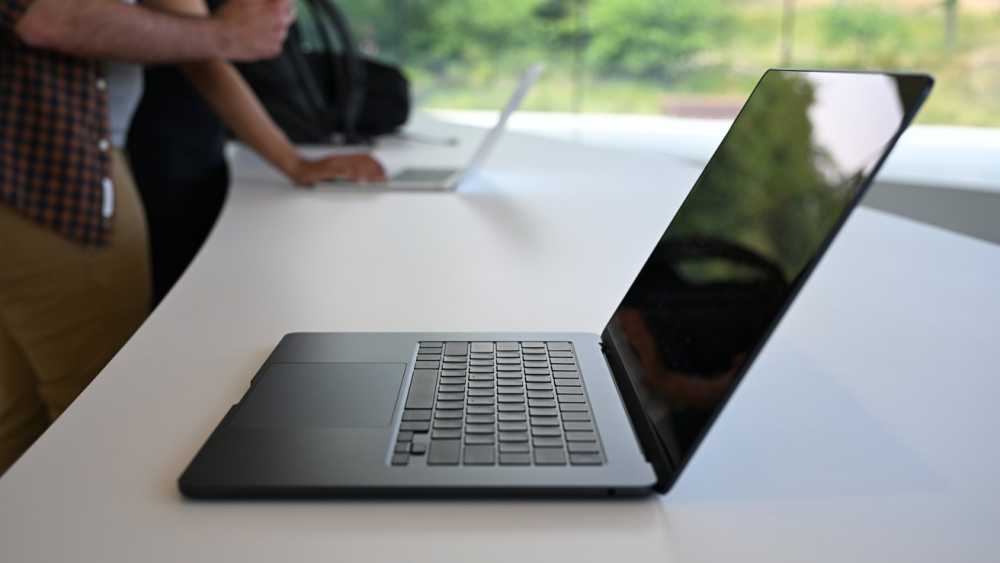 Open laptop on white table, with another person using a laptop in the background, against a large window with greenery outside.