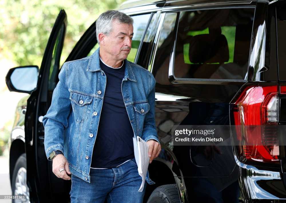 Man wearing denim jacket and jeans steps out of a black SUV, holding papers in one hand.