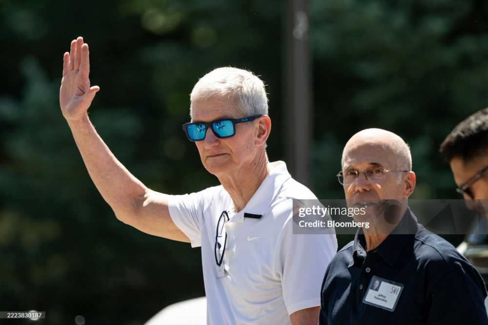 Man in white shirt and sunglasses waving, standing next to another man with glasses wearing a dark shirt. Trees are blurred in the background.