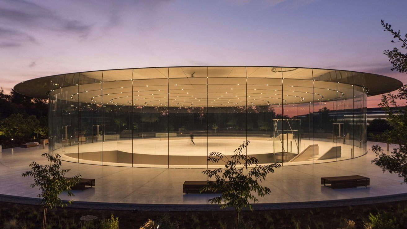 Modern circular glass building with a large flat roof at sunset, surrounded by trees and benches.