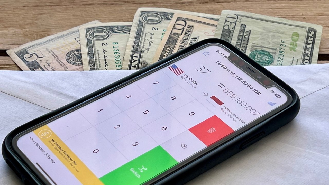Smartphone with currency converter app displayed, showing USD to IDR conversion. Background includes a five-dollar bill, two one-dollar bills, and a twenty-dollar bill on a wooden surface.